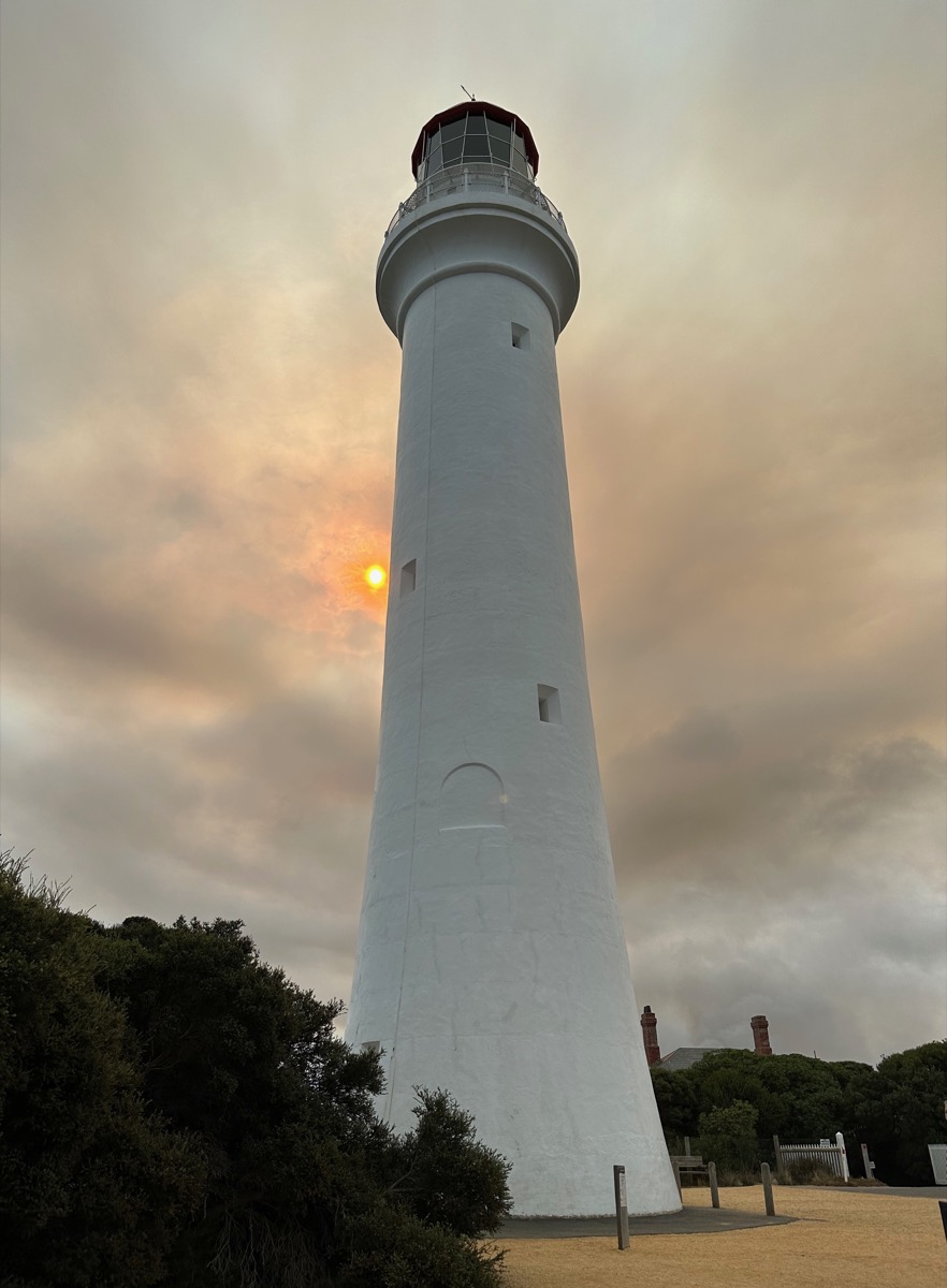 White lighthouse with red top under cloudy sky; sun partially visible.